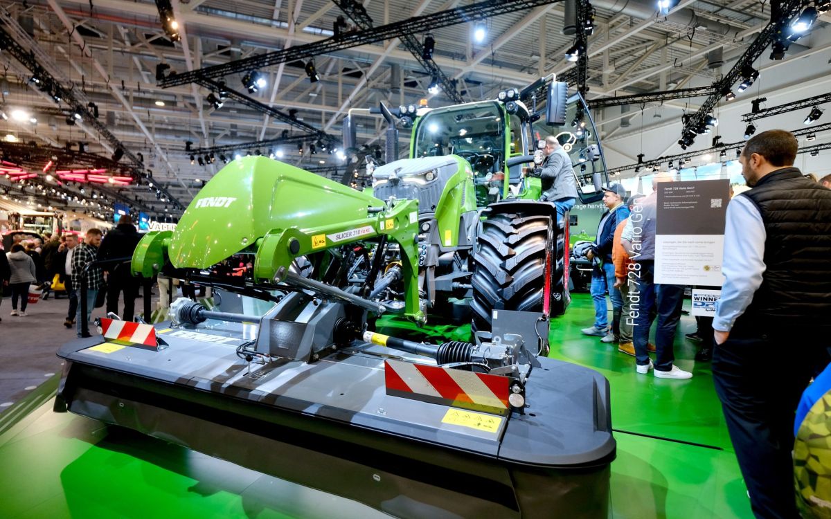 Un grand tracteur Fendt vert avec une faucheuse frontale intégrée se tient dans un hall d’exposition. Autour de lui, des visiteurs observent la machine et l’exposition.